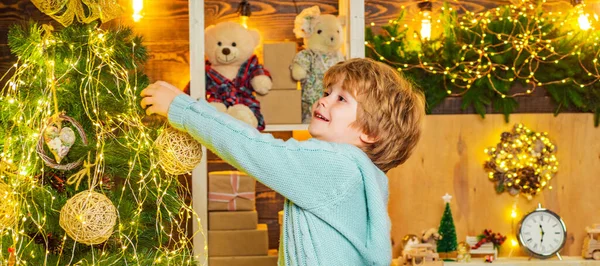 Cute kid decorating Christmas tree with bauble. Christmas preparation funny little boy celebrating New Year. Child is decorating the Christmas tree with Christmas ball