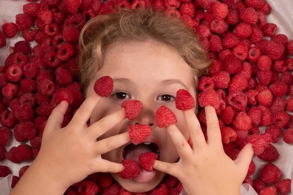 Funny kids face near raspberry background. Cute child eats raspberries from fingers. Kids face in raspberries fruits, healthy kids nutrition concept