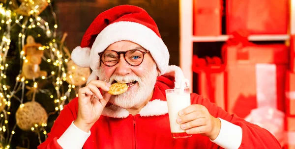 サンタは面白い顔をしてクッキーを選ぶ 熟女サンタはクッキーを食べ 自宅で牛乳のガラスを保持します メリークリスマスと幸せな新年 — ストック写真