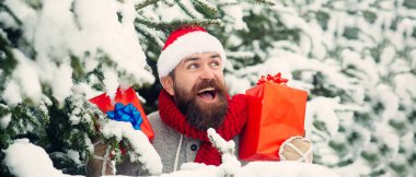 Noel Baba kışın karlı arka planda hediye kutusuyla heyecanlanıyor. Noel Baba şapkalı, açık havada Noel hediyeli bir adam. Karlı kış arkaplanı. Noel Baba dışarıda kar yağışı altında hediyeler taşıyor.