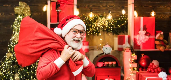 白い髭を生やしたサンタの祖父はクリスマスの木製の背景にポーズをとったプレゼント付きのバッグを持っています サンタクロースとクリスマスグリーティングカード — ストック写真