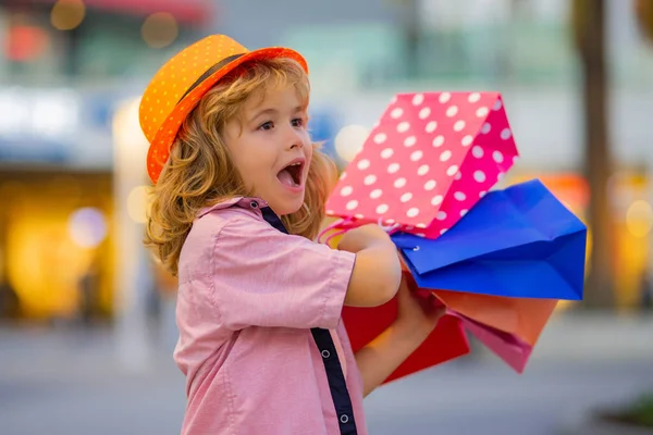 Excited shopper. Kid with shopping bags. Child in trendy hat and shirt shopping near shopping center. Happy boy holding shopping bags at the mall