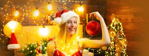 美しい若い女性サンタはクリスマスの装飾されたインテリアの背景にクリスマスボールを保持します クリスマスの光の中で赤い唇を持つ笑顔の女性 クリスマスの女の子気分 — ストック写真