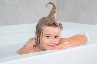 Şampuan köpüğü ve banyo yapan saçlarında kabarcıklar olan bir çocuk. Köpüklü çocuk suratı, saç bakımı ve hijyen konsepti. Sabun köpüklü banyo küveti