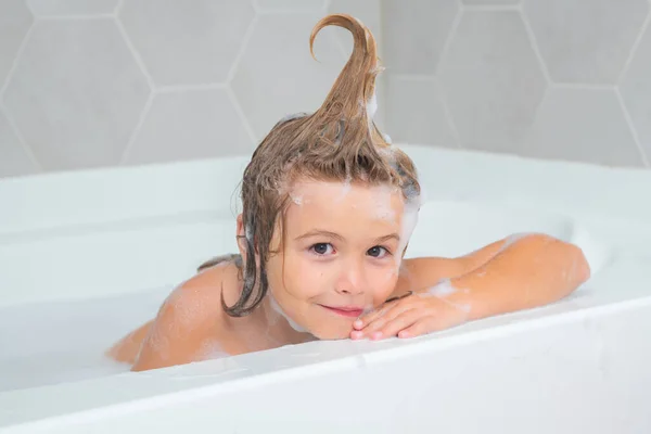 Child with shampoo foam and bubbles on hair taking bath. Funny face of kid in foam, hair care and hygiene concept. Bath tub with soap bubble