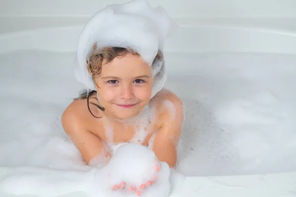 Kids face in foam. Child boy washing with a bubbles in bath. Cute child bathes, lying in a white bath with foam from soap and shampoo