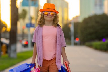 Moda çocuğu alışverişte. Alışveriş çantalı bir çocuğun portresi. Tişörtlü, güneş gözlüklü ve şapka alışverişi yapan moda çocuğu. Sokaklarda alışveriş torbaları tutan mutlu çocuk.