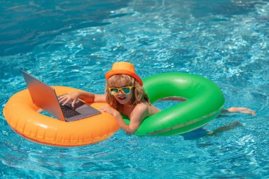 Yaz ofisi ve serbest çalışma konsepti. Çocuk havuzda yüzme ringinde oturuyor ve dizüstü bilgisayar kullanıyor. İnternetten alışveriş, serbest konsept, yaz seyahati.