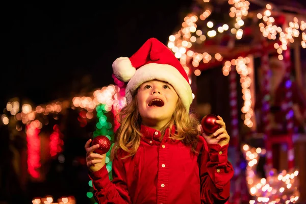 Christmas decoration in front of a night house. Happy kid looks at the sky in the Christmas night. Kid in santa hat near home garlands outside. House Decorated with Garlands