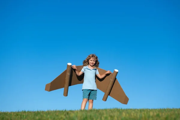 Child boy plays in an astronaut dreams of space. Happy child play with toy plane cardboard wings against blue sky. Kid having fun in summer field outdoor. Portrait of boy with paper wings