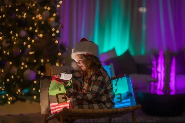 Kid with present gift with magic light. Lighting present gift bag. Happy child in pajamas near Christmas tree at home with traditional Christmas tree