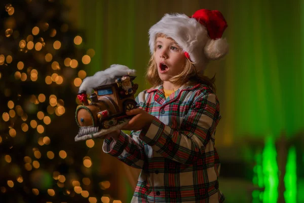 Child celebrating Christmas at home. Kid in santa hat on front of night Christmas tree home background on Christmas Eve. Happy child in checkered Christmas pajama enjoying holiday evening at home