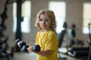 Spor salonundaki çocuk. Çocuklarla boş zamanlarında spor aktiviteleri. Dambıl tutan sarışın çocuk. Çocuklar için spor egzersizleri. Halterleri kaldıran komik çocuk.
