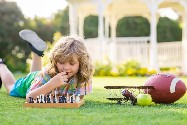 Summer games and outdoor activities for kids. Child playing chess game in spring backyard, laying on grass. Concentrated kid play chess. Kid playing board game outdoor