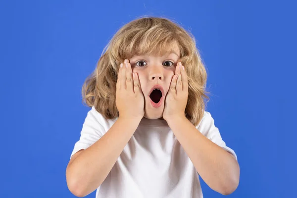 Shocked kid boy keeping hands near cheek with open mouth on blue isolated background. Surprised face, excited emotions of child. Shock, omg and wow expression