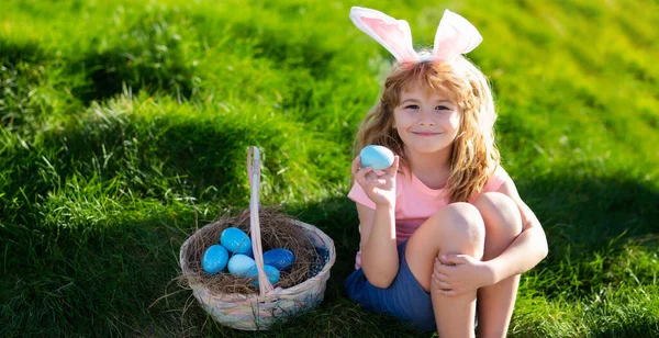 Horizontal photo banner for website header design. Happy Easter for children. Boy in bunny ears with colorful eggs play and hunting easter eggs outside. Cute child with easter basket on grass