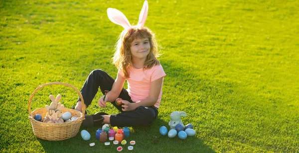 Child with easter eggs and bunny ears laying on grass. Happy Easter kids face. Cute little boy, easter bunny children spring outdoor. Panoramic web banner frame