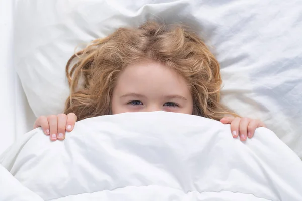 Kid under covers, face cover with blanket. Child wakes up in the morning in the bedroom. Cute little boy waking up in bed