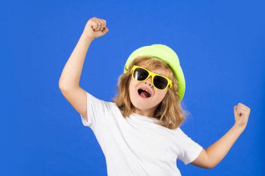 Mavi stüdyonun arka planında yaz şapkalı, tişörtlü ve güneş gözlüklü çocuk portresi. Stüdyodaki heyecanlı çocuk izole edilmiş. Şaşkın yüz, şaşkın çocuk duyguları.