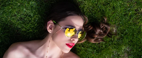 Spring woman. Girl in sunglasses enjoying nature lying on grass. Beauty trends. Copyspace. Summer reflection in sunglasses of beautiful young woman on grass. Banner for website header