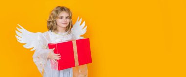 Cute blonde kid angel with gift box present. Child angel. Portrait of cute kid with angel wings isolated on yellow studio background. Horizontal header, banner