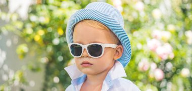 Close up face of cute child outdoors. Spring banner for website header. Kids fashion. Happy child in sunglasses and hat, adorable lovely kid. Joyful portrait of small child on green nature.