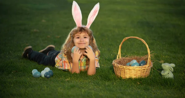 Child boy hunting easter eggs in spring lawn laying on grass. Bunny kids with rabbit bunny ears