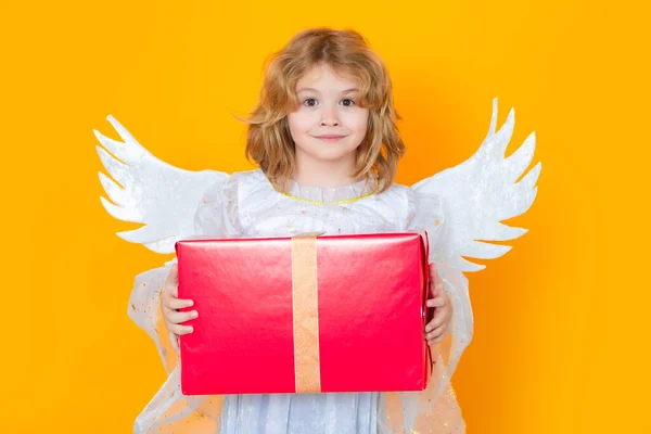 Cute blonde kid angel with gift box present. Valentine angel. Little cupid child. Kid angel with angels wings, isolated on yellow studio background. Valentines day gift card