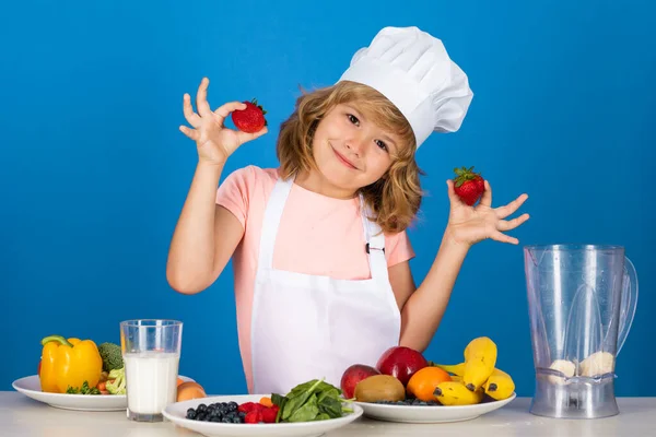 Child wearing cooker uniform and chef hat preparing vegetables on kitchen hold strawberries, studio portrait. Cooking, culinary and kids food concept