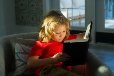 Portrait of cute blonde child reading interesting kids book story. Child reading book at home. Little boy sitting on couch in living room reading story book. Kid doing homework for elementary school