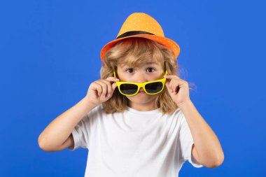 Fashion portrait of amazed kid in summer hat, t-shirt and sunglasses on blue studio isolated background