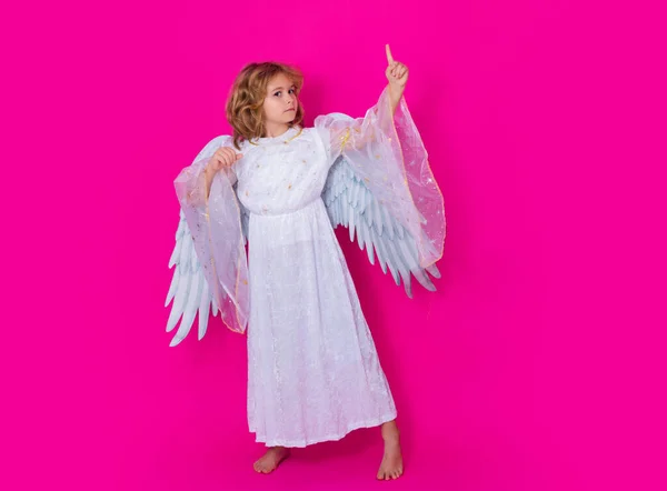 Child angel pointing up, point gesture. Valentines day. Blonde cute child with angel wings on a pink red studio background. Happy angel child