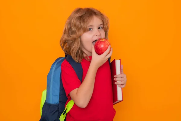 School kids eat apple. Child from elementary school with book and apple isolated on yellow background. Little student, clever nerd pupil ready to study. Knowledge, education and learning