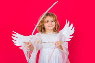 Angel kid shoots a love arrow from a bow on Valentines Day. Valentines day. Blonde cute child with angel wings on a red studio background. Happy angel child