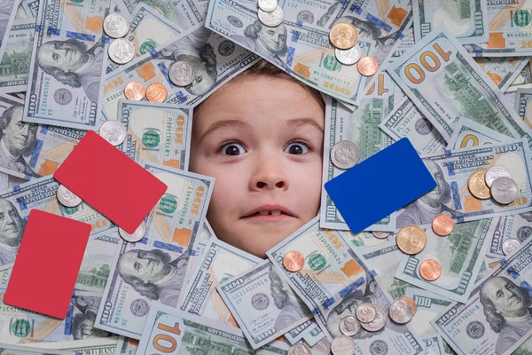 Funny child with fun face with money. Kid peeking out of dollar bills with astonished shocked eyes. Money background. Shopping and financial concept. Cash dollars banknotes, coins and credit card