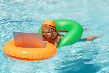 Child relaxing in the pool with laptop. Kid online working on laptop, swimming in a sunny turquoise water pool. Summer office and freelance concept