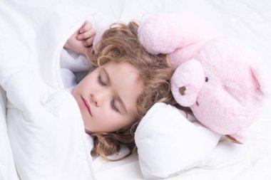 Little boy sleeps. Lovely face of blonde caucasian child, sleeping with a toy teddy bear on bed. Sweet dreams. Little baby boy sleeping while lying on bed at home