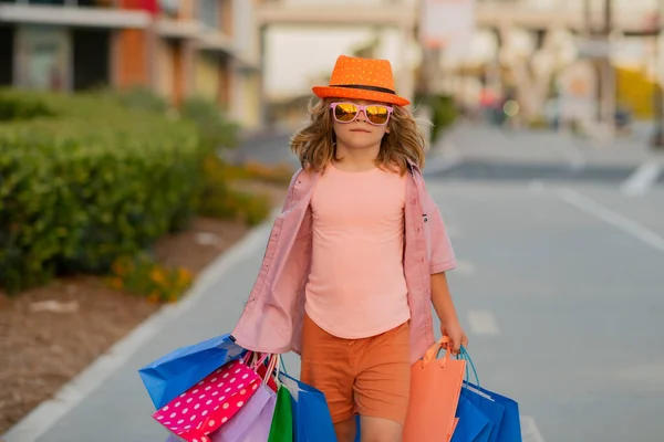 Shopper. Cute little boy in summer fashion clothes goes shopping. Happy child with shopping packages in hands. Shopper child with shopping bag. Cute kid walking on street carrying shopping bags