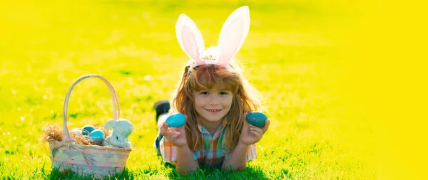 Child boy hunting easter eggs. Wide photo banner for website header design. Kid boy lying on the grass and finding easter eggs. Child boy with easter eggs and bunny ears on grass