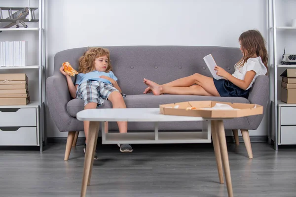 Home kids leisure. Entertainment for children. Cute kids eating pizza reading book enjoying leisure at home. Two little children relaxing at home. Children friends spend time together in living room