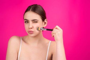 Utangaç bir kadın stüdyo arka planında izole edilmiş toz yüzü uygular. Yanaklarını pudralayan kız. Makyaj fırçası. Kadın modelin elmacık kemikleri kızarır.