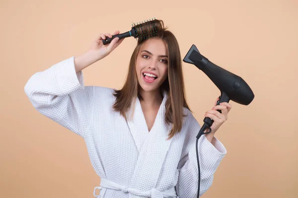 Woman in bathrobe combing hair, drying hairs with hairdryer. Portrait of female model with a comb brushing hair. Girl with hair brush and blow dryer. Hair care and beauty. Morning routine