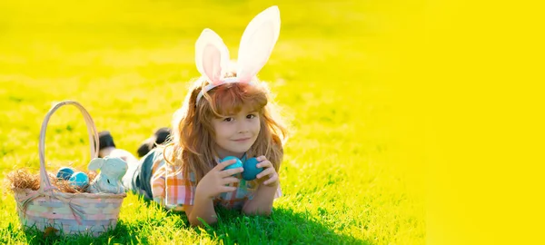 Wide photo banner for website header design. Child boy laying on grass hunting easter eggs. Cute kid in rabbit costume with bunny ears having easter in park. Children hunt easters egg