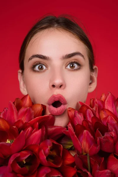 Surprised woman face with flowers. Beautiful sensual woman hold tulips bouquet, studio portrait on red background. Close up beauty woman face near tulips bouquet