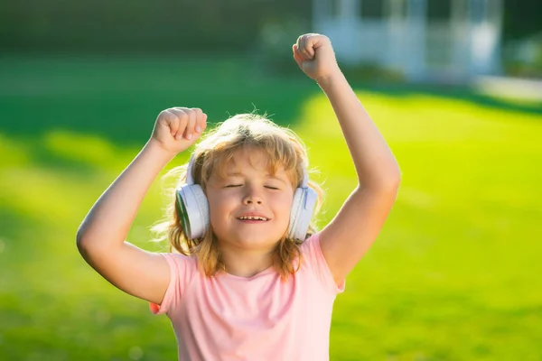 Happy child enjoys listens to music in headphones over green grass background. Funny kid in headphones listening to music on summer park or backyard outdoor