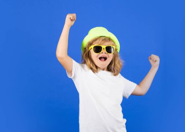 Excited kid boy on studio isolated background. Fashion portrait of kid in summer hat, t-shirt and sunglasses on blue studio isolated background. Surprised face, amazed emotions of child