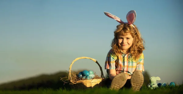Wide photo banner for website header design. Cute bunny child boy with rabbit ears. Children hunting easter eggs on sky background with copy space