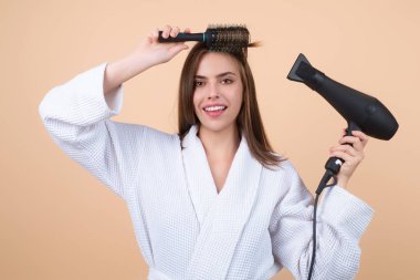 Bornozlu kadın saç tarıyor, saç kurutma makinesiyle saç kurutuyor. Saçlarını tarayan kadın modelin portresi. Saç fırçası ve saç kurutma makinesi olan kız. Saç bakımı ve güzellik. Sabah rutini