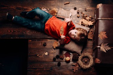 Çocuklar için tüm sonbahar kıyafetlerinde en büyük indirim. Sarışın, küçük bir çocuk, sonbahar yapraklarında, midesinde yaprak, ahşap zeminde yatıyor. Sonbaharda oynayan çocuk