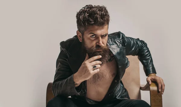Brutal bearded fashionable man with serious face sitting in chair and looking at camera. Macho man Casanova attracting women. Sexy rich shirtless millionaire man with leather jacket
